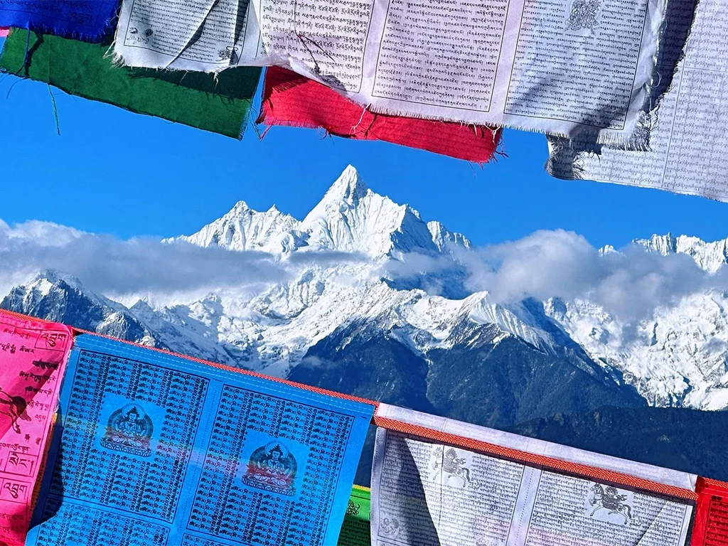 Diqing Meili Snow Mountain 迪慶梅里雪山壯麗雪峰景觀,潔白山巒在藍天與雲霧間聳立,前景飄揚的五彩經幡增添濃厚藏族宗教氛圍,展現梅里雪山神聖與壯闊的自然景色,是迪慶香格里拉地區著名的高原旅遊與朝聖地標