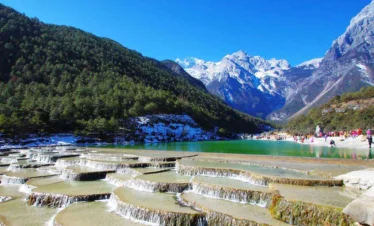 玉龍雪山白水台呈現層層堆疊的白色鈣化梯田景觀，清澈流水沿著階梯狀平台緩緩流下。遠方可見雪覆的玉龍雪山與翠綠山谷，藍天映襯壯麗自然景色，遊客在湖畔與平台附近欣賞玉龍雪山白水台獨特地貌。