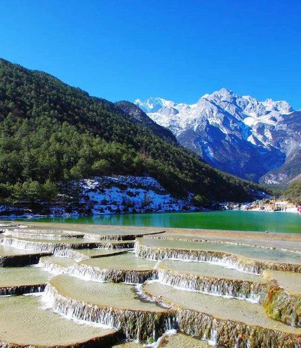 玉龍雪山白水台呈現層層堆疊的白色鈣化梯田景觀，清澈流水沿著階梯狀平台緩緩流下。遠方可見雪覆的玉龍雪山與翠綠山谷，藍天映襯壯麗自然景色，遊客在湖畔與平台附近欣賞玉龍雪山白水台獨特地貌。