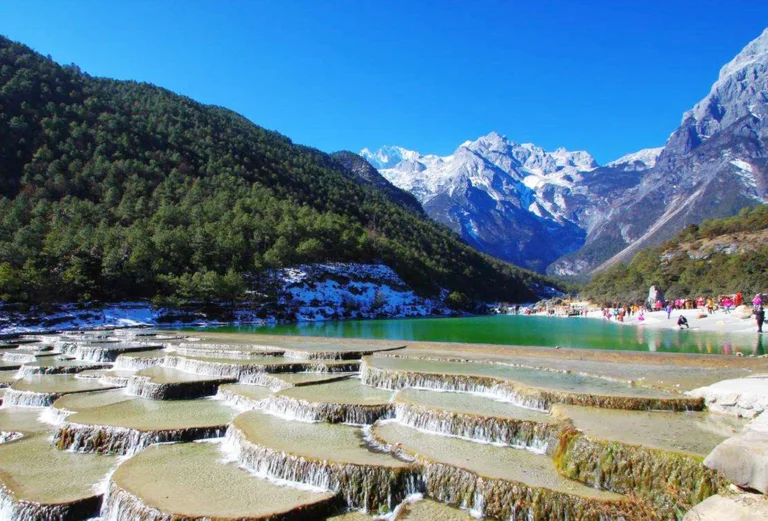 玉龍雪山白水台呈現層層堆疊的白色鈣化梯田景觀，清澈流水沿著階梯狀平台緩緩流下。遠方可見雪覆的玉龍雪山與翠綠山谷，藍天映襯壯麗自然景色，遊客在湖畔與平台附近欣賞玉龍雪山白水台獨特地貌。