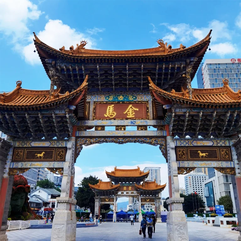 昆明金馬碧雞坊矗立於廣場，藍天下的傳統建築展現飛簷翹角與精美彩繪。牌坊刻有「金馬」字樣，與後方現代城市建築交織。