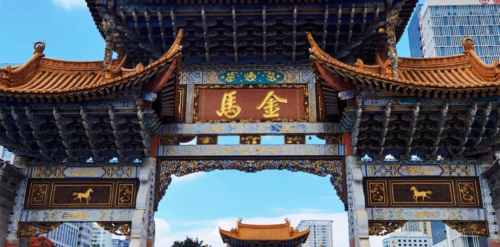 昆明金馬碧雞坊矗立於廣場，藍天下的傳統建築展現飛簷翹角與精美彩繪。牌坊刻有「金馬」字樣，與後方現代城市建築交織。