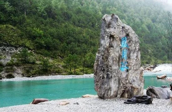麗江藍月谷湖水呈現清澈的藍綠色，河谷被翠綠山林環繞，岸邊立著刻有「藍月谷」字樣的天然巨石，湖水與森林相互映襯，展現玉龍雪山腳下寧靜而夢幻的自然風景。
