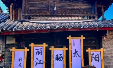 麗江古城傳統木造建築外觀，屋頂覆蓋灰色瓦片，展現納西族古城建築特色。門前擺放花卉盆栽與木製展示架，上方書寫「我在麗江晒太陽」字樣，呈現悠閒的古城生活氛圍，是遊客拍照與體驗麗江古城文化的熱門場景。