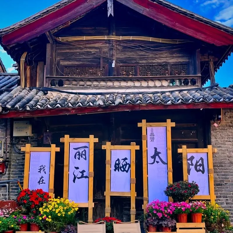 麗江古城傳統木造建築外觀，屋頂覆蓋灰色瓦片，展現納西族古城建築特色。門前擺放花卉盆栽與木製展示架，上方書寫「我在麗江晒太陽」字樣，呈現悠閒的古城生活氛圍，是遊客拍照與體驗麗江古城文化的熱門場景。