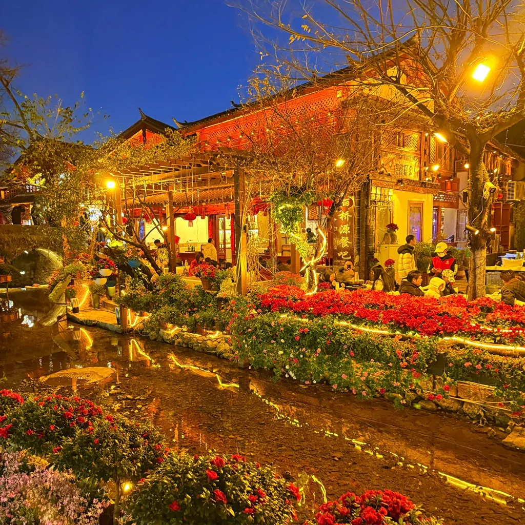 夜晚的 麗江古城 燈火通明，小橋流水與花卉點綴其間，街邊餐廳與遊客交織，營造出溫馨浪漫的古城氛圍。