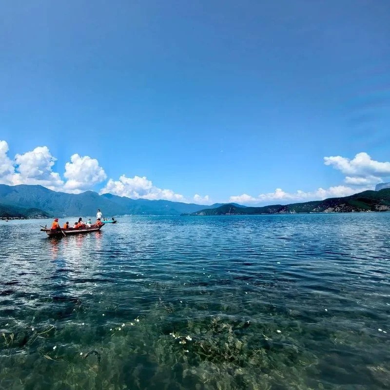 Lugu Lake pig trough boat 晴朗藍天下的瀘沽湖湖面清澈平靜,遠方群山層疊延伸至天際,湖水透出翠綠與湛藍色澤。前景可見一艘載著遊客的小船在湖面緩緩前行,水下石頭與水草清晰可見,展現瀘沽湖純淨自然與寧靜壯麗的高原湖泊風光。