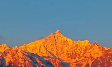 清晨陽光照耀 梅里雪山，雪峰被金色光芒染亮，山體輪廓清晰壯麗，天空湛藍，呈現震撼的「日照金山」自然奇觀。