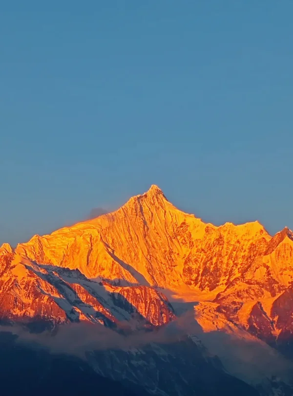 清晨陽光照耀 梅里雪山，雪峰被金色光芒染亮，山體輪廓清晰壯麗，天空湛藍，呈現震撼的「日照金山」自然奇觀。