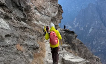 在 虎跳峡 悬崖栈道上徒步的游客，背着登山包沿着崎岖山路前行，峡谷深邃壮观，远处山峰连绵，展现惊险与壮美并存的自然风光。