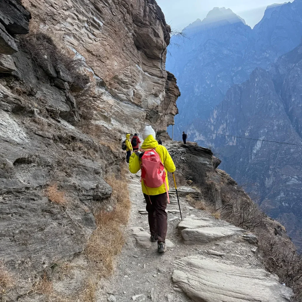 在 虎跳峡 悬崖栈道上徒步的游客，背着登山包沿着崎岖山路前行，峡谷深邃壮观，远处山峰连绵，展现惊险与壮美并存的自然风光。