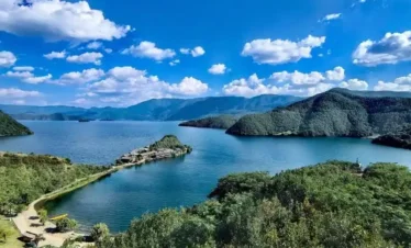 瀘沽湖壯麗湖景在晴朗藍天下展開，湖水深藍清澈，群山環繞湖泊。湖中央的小島與連接岸邊的堤道清晰可見，周圍綠意盎然的森林與遠方山巒層層延伸，白雲點綴天空，展現瀘沽湖高原湖泊寧靜而壯觀的自然風光。
