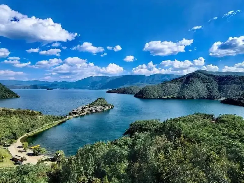 瀘沽湖壯麗湖景在晴朗藍天下展開，湖水深藍清澈，群山環繞湖泊。湖中央的小島與連接岸邊的堤道清晰可見，周圍綠意盎然的森林與遠方山巒層層延伸，白雲點綴天空，展現瀘沽湖高原湖泊寧靜而壯觀的自然風光。