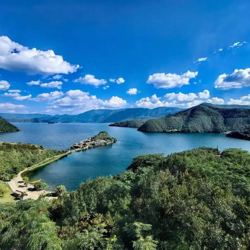瀘沽湖壯麗湖景在晴朗藍天下展開，湖水深藍清澈，群山環繞湖泊。湖中央的小島與連接岸邊的堤道清晰可見，周圍綠意盎然的森林與遠方山巒層層延伸，白雲點綴天空，展現瀘沽湖高原湖泊寧靜而壯觀的自然風光。