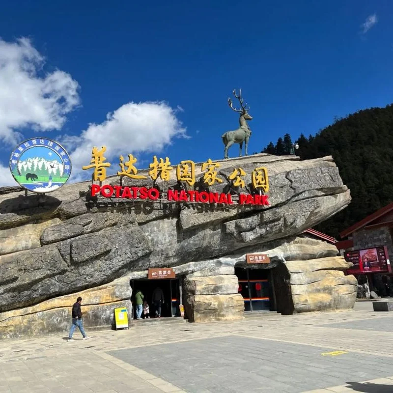 Pudacuo National Park Shangri La 香格里拉普達措公園入口以巨型岩石造型打造,上方設有鹿雕像與「普達措國家公園」標誌字樣。藍天白雲與森林山景環繞,遊客從岩石洞口入口進入景區,呈現香格里拉普達措公園自然與生態特色的代表性地標景觀。