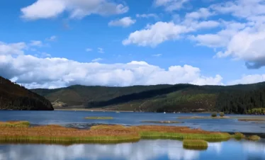位于 属都湖 的高原湖泊景观，湖水清澈平静，周围草甸与森林环绕，远山起伏，蓝天白云倒映水中，风光宁静优美。