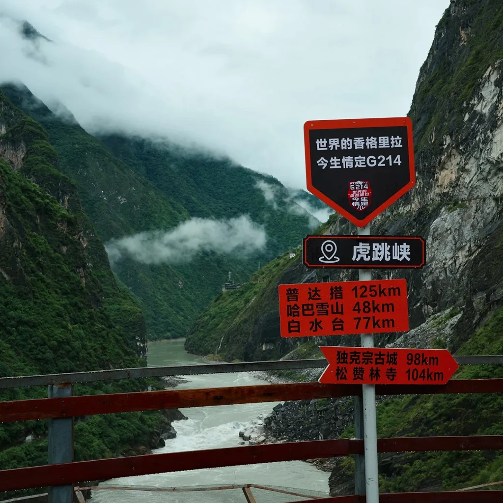 壯麗的 香格里拉虎跳峽 景色，翠綠峽谷被雲霧環繞，下方金沙江奔流，前方設有觀景路牌。