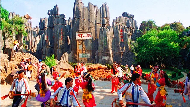stone forest 1 昆明石林景區內高聳的石灰岩石峰形成壯觀的喀斯特地貌,前方廣場上少數民族表演團隊身穿鮮豔傳統服飾載歌載舞。遊客在周圍觀賞拍照,背景可見刻有「石林」字樣的石壁,呈現昆明石林自然景觀與民族文化活動。