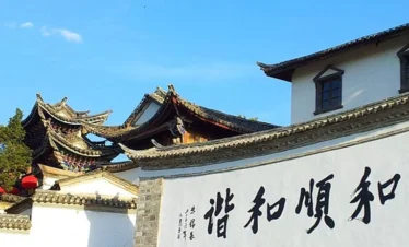 雲南騰衝的 和順古鎮 古建築景觀。白色石牆上寫著「和順和諧」書法字樣，牆後可見傳統中式屋頂與飛簷建築，青瓦層疊，展現典型的古鎮風格。晴朗藍天下，古鎮街巷寧靜雅致，整體畫面呈現濃厚的歷史文化氣息與雲南傳統人文風貌。