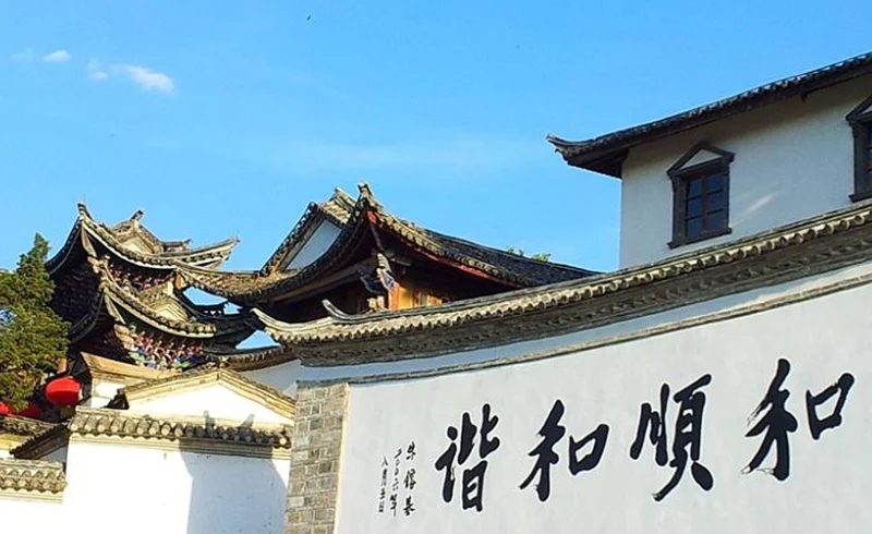 雲南騰衝的 和順古鎮 古建築景觀。白色石牆上寫著「和順和諧」書法字樣，牆後可見傳統中式屋頂與飛簷建築，青瓦層疊，展現典型的古鎮風格。晴朗藍天下，古鎮街巷寧靜雅致，整體畫面呈現濃厚的歷史文化氣息與雲南傳統人文風貌。