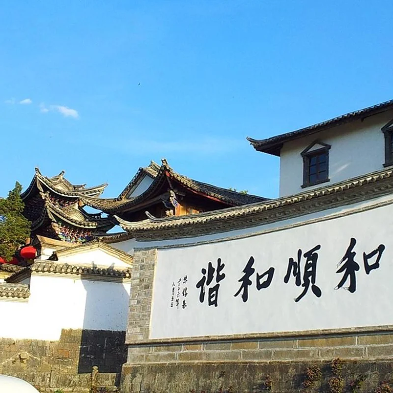雲南騰衝的 和順古鎮 古建築景觀。白色石牆上寫著「和順和諧」書法字樣，牆後可見傳統中式屋頂與飛簷建築，青瓦層疊，展現典型的古鎮風格。晴朗藍天下，古鎮街巷寧靜雅致，整體畫面呈現濃厚的歷史文化氣息與雲南傳統人文風貌。