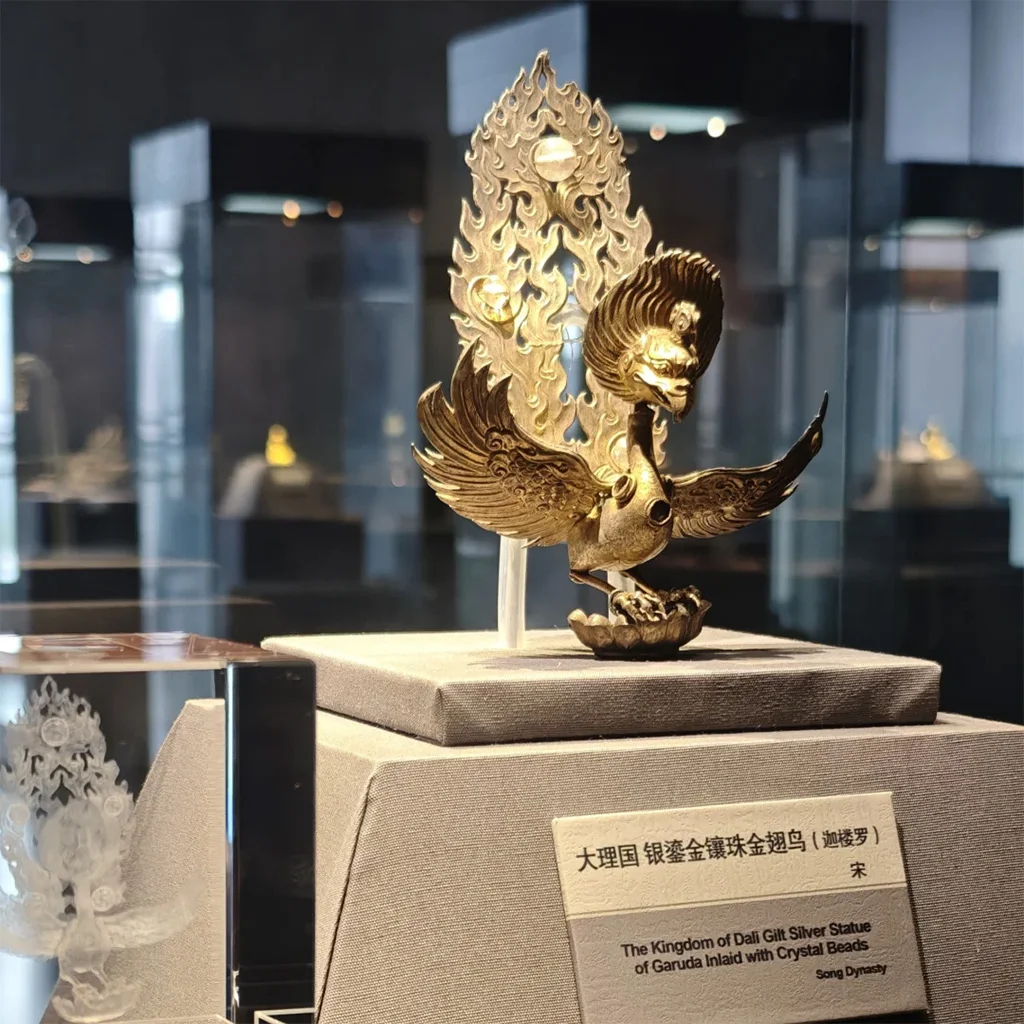 於 雲南國家寶藏館 展廳中，一件金色迦樓羅造型文物展翅而立，背後火焰紋裝飾華麗，細節精緻，展現濃厚宗教藝術風格。