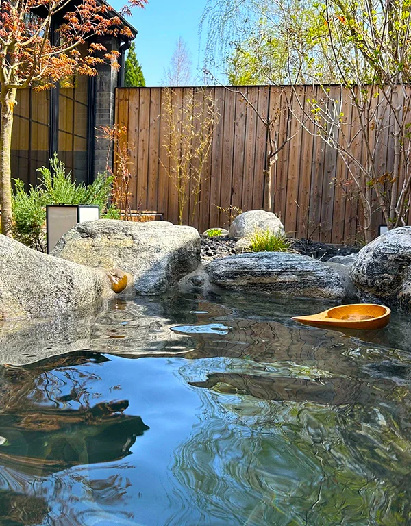 騰衝溫泉庭園露天湯池景觀，清澈溫泉水環繞天然岩石與日式庭園造景，木牆與綠樹點綴寧靜環境，木製湯勺漂浮水面，呈現騰衝溫泉放鬆療癒的泡湯氛圍，是雲南著名的溫泉養生旅遊景點。