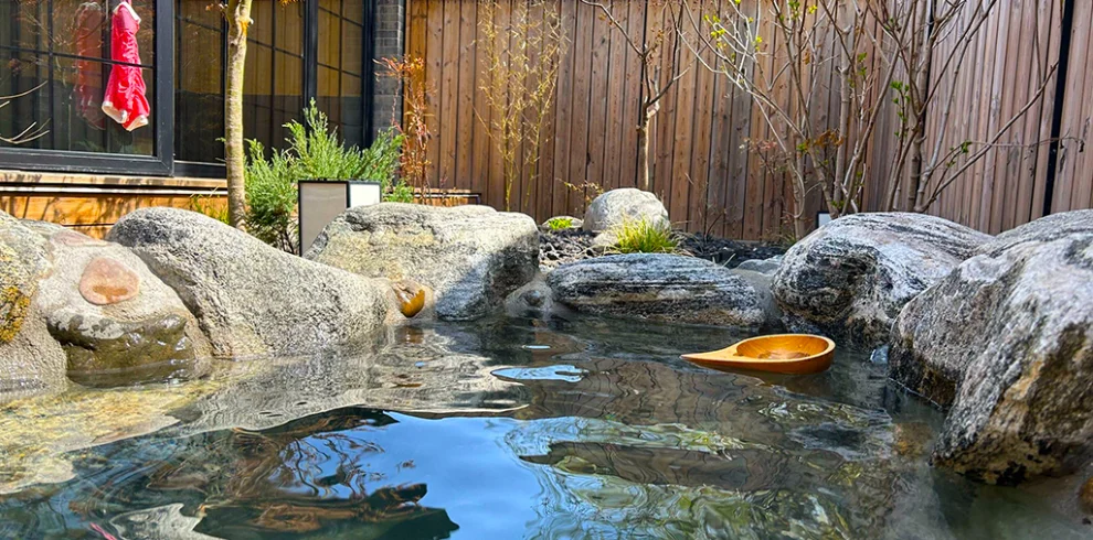 騰衝溫泉庭園露天湯池景觀，清澈溫泉水環繞天然岩石與日式庭園造景，木牆與綠樹點綴寧靜環境，木製湯勺漂浮水面，呈現騰衝溫泉放鬆療癒的泡湯氛圍，是雲南著名的溫泉養生旅遊景點。