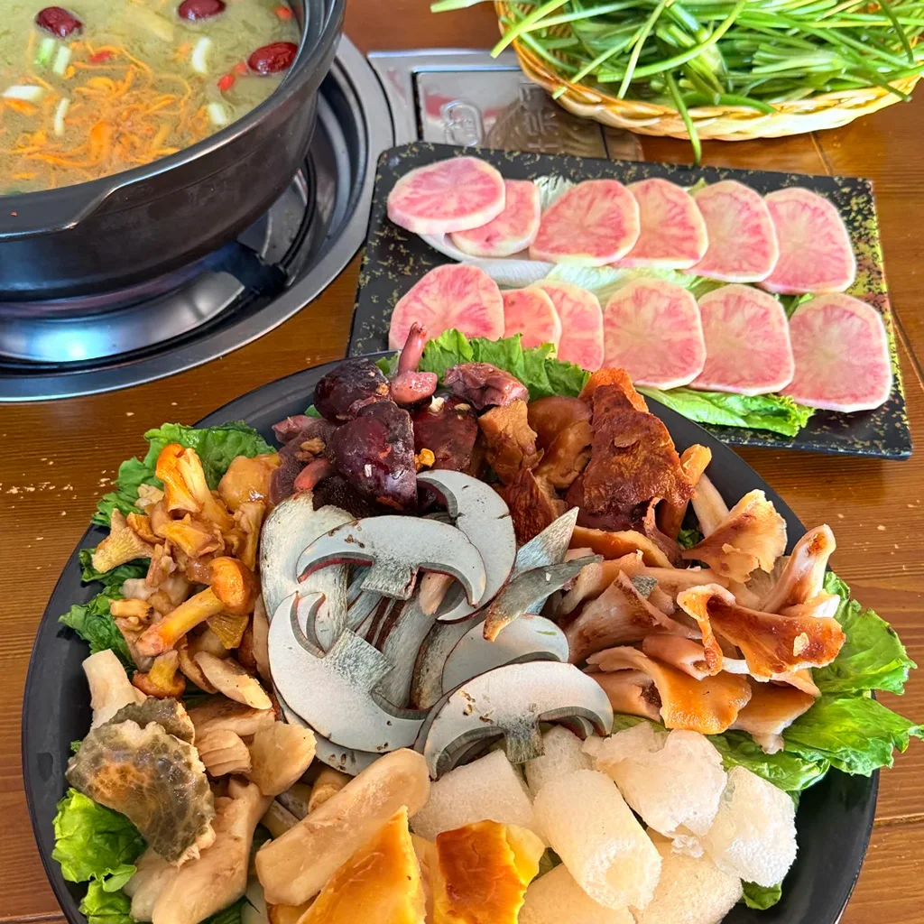 黑色大盤中擺放著多種新鮮野生菌切片，配上一鍋沸騰的高湯與粉紅色蘿蔔片，展現道地雲南風味。