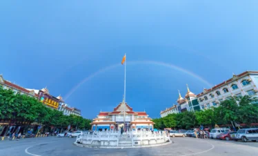 姐告口岸廣場景觀，中央設有圓形噴泉與高聳旗桿，周圍為具有東南亞風格的建築群與商店街。天空出現一道完整彩虹橫跨廣場上方，與藍天形成壯麗畫面，展現雲南邊境城市熱鬧而獨特的口岸景觀與旅遊氛圍。