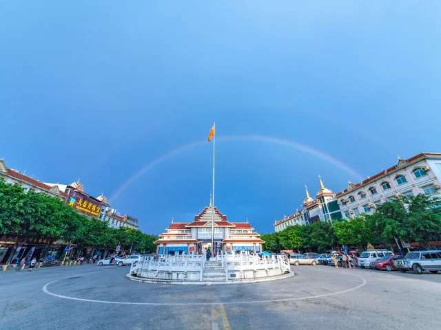 姐告口岸廣場景觀，中央設有圓形噴泉與高聳旗桿，周圍為具有東南亞風格的建築群與商店街。天空出現一道完整彩虹橫跨廣場上方，與藍天形成壯麗畫面，展現雲南邊境城市熱鬧而獨特的口岸景觀與旅遊氛圍。