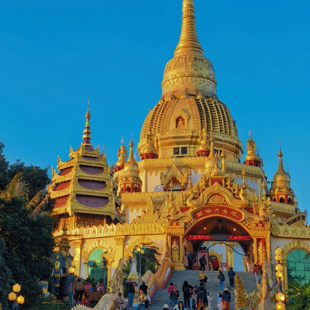 Meng Huan Grand Golden Pagoda2 中國雲南芒市勐煥大金塔,宏偉鍍金佛塔在夕陽下閃耀,遊客沿著宏偉階梯參觀,背景是澄澈藍天。