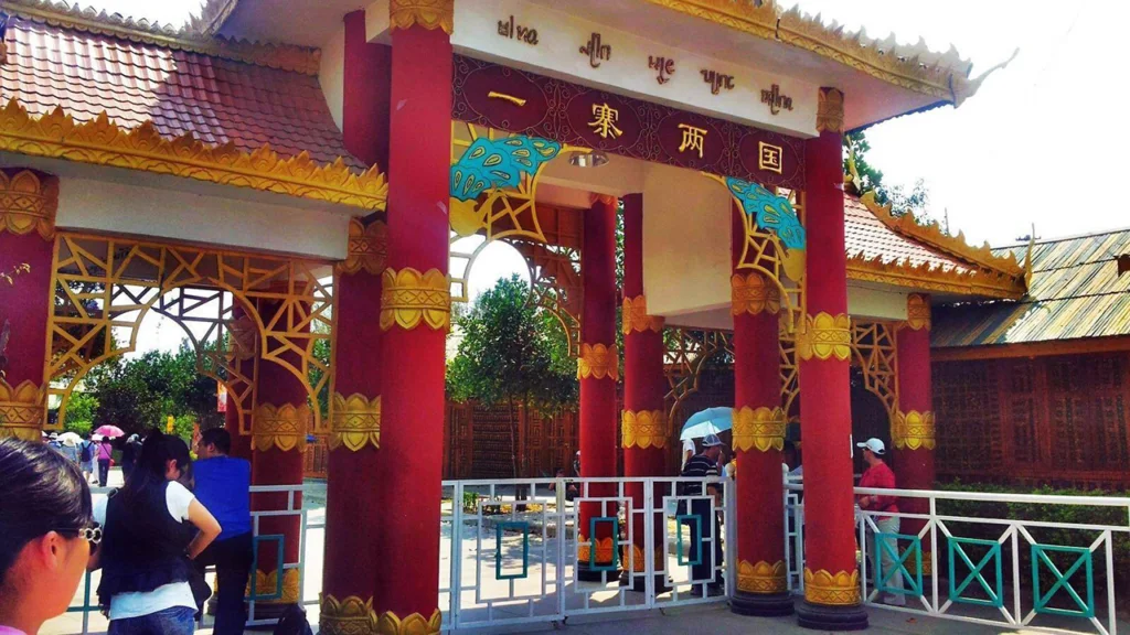 一寨兩國景區入口建築，紅色立柱與金色裝飾呈現東南亞民族風格，上方牌匾寫著「一寨兩國」，遊客在入口處參觀拍照，展現中緬邊境一寨兩國獨特的地理與文化景觀。