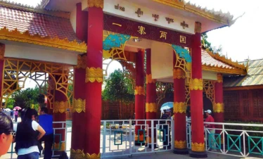 一寨兩國景區入口建築，紅色立柱與金色裝飾呈現東南亞民族風格，上方牌匾寫著「一寨兩國」，遊客在入口處參觀拍照，展現中緬邊境一寨兩國獨特的地理與文化景觀。