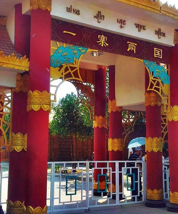 一寨兩國景區入口建築，紅色立柱與金色裝飾呈現東南亞民族風格，上方牌匾寫著「一寨兩國」，遊客在入口處參觀拍照，展現中緬邊境一寨兩國獨特的地理與文化景觀。