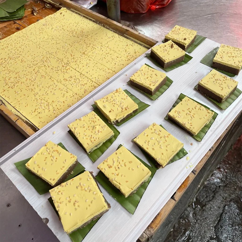 Osmanthus cake 地道騰衝桂花蛋糕,雲南特色甜點。金黃色方形糕點灑滿芝麻,內餡夾層為紅豆沙,置於芭蕉葉上,展現傳統在地手藝