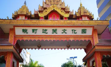 萬慶邊關文化公園入口牌樓，金紅色緬甸風格屋頂與精緻雕飾十分醒目，門樓上方寫有「萬慶邊關文化園」，象徵中緬邊境文化交流特色，入口道路兩側綠樹成蔭與停放車輛，展現邊境城市獨特的人文景觀與旅遊氛圍。