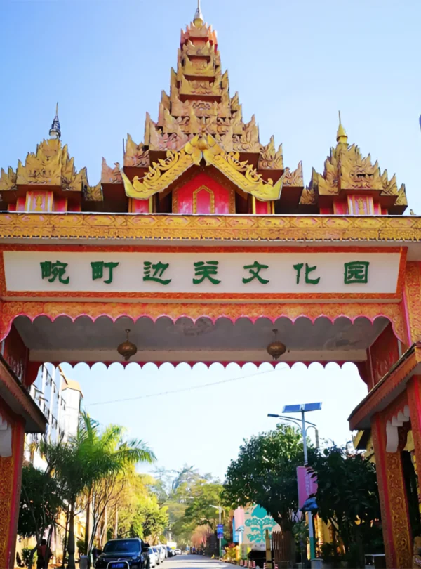 萬慶邊關文化公園入口牌樓，金紅色緬甸風格屋頂與精緻雕飾十分醒目，門樓上方寫有「萬慶邊關文化園」，象徵中緬邊境文化交流特色，入口道路兩側綠樹成蔭與停放車輛，展現邊境城市獨特的人文景觀與旅遊氛圍。
