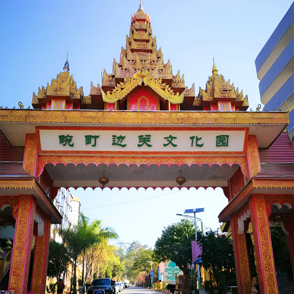 萬慶邊關文化公園入口牌樓，金紅色緬甸風格屋頂與精緻雕飾十分醒目，門樓上方寫有「萬慶邊關文化園」，象徵中緬邊境文化交流特色，入口道路兩側綠樹成蔭與停放車輛，展現邊境城市獨特的人文景觀與旅遊氛圍。