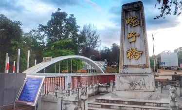 萬慶橋入口景觀，橋前立有刻著「萬慶橋」的石碑，旁邊是白色拱形橋體與紅色欄杆設計，周圍綠樹環繞與步道景觀，呈現邊境小鎮寧靜氛圍。此橋為當地具有歷史意義的地標之一，也是遊客參觀與拍照的熱門景點。