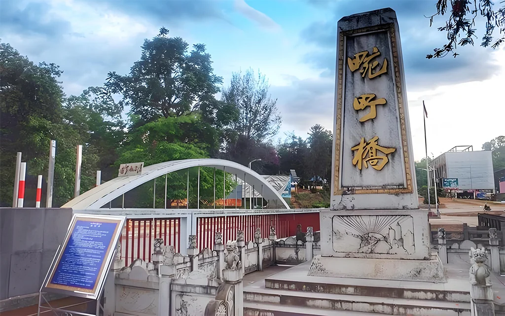 萬慶橋入口景觀，橋前立有刻著「萬慶橋」的石碑，旁邊是白色拱形橋體與紅色欄杆設計，周圍綠樹環繞與步道景觀，呈現邊境小鎮寧靜氛圍。此橋為當地具有歷史意義的地標之一，也是遊客參觀與拍照的熱門景點。