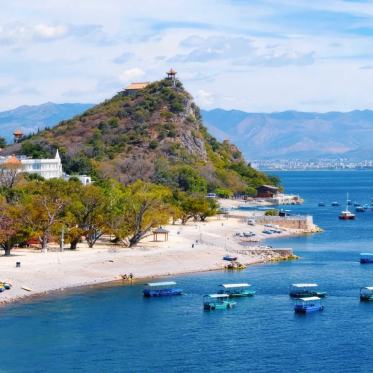 雲南撫仙湖湖畔風景，湛藍湖水與沙灘停泊的小船形成寧靜畫面，山丘頂端的古亭與寺閣俯瞰整片湖面，周圍綠樹與遠山層層延伸，展現撫仙湖清澈遼闊的高原湖泊景觀與悠閒度假氛圍。
