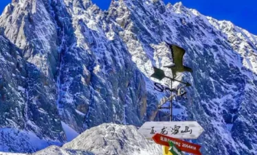 雲南麗江著名的 玉龍雪山 壯麗雪峰景觀。高聳陡峭的雪山覆蓋著白雪，在湛藍天空下顯得格外壯觀。前景是一個寫著「玉龍雪山」的方向指示牌與風向標，指向世界多個城市距離，為畫面增添旅遊地標特色，展現出這座著名雪山的雄偉與魅力。