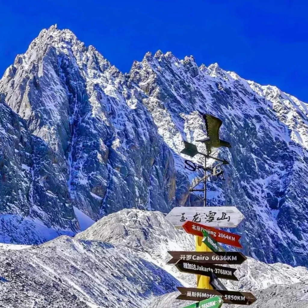 雲南麗江著名的 玉龍雪山 壯麗雪峰景觀。高聳陡峭的雪山覆蓋著白雪，在湛藍天空下顯得格外壯觀。前景是一個寫著「玉龍雪山」的方向指示牌與風向標，指向世界多個城市距離，為畫面增添旅遊地標特色，展現出這座著名雪山的雄偉與魅力。