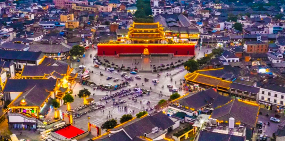 雲南建水古城黃昏城市景觀，空拍視角可見城門與古建築在燈光點綴下格外醒目，街道向四方延伸，周圍傳統屋頂與現代城市交織，展現建水古城歷史文化與熱鬧生活交融的景象。