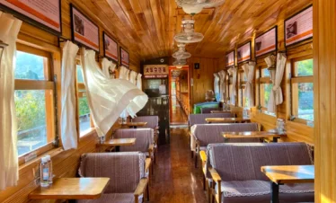 建水小火車復古車廂內部景觀，木質天花板與牆面營造溫暖氛圍，兩側窗戶配有白色窗簾與木桌座椅，車廂內整齊排列的座位與吊扇呈現懷舊鐵道風格，展現建水小火車獨特的歷史與觀光魅力