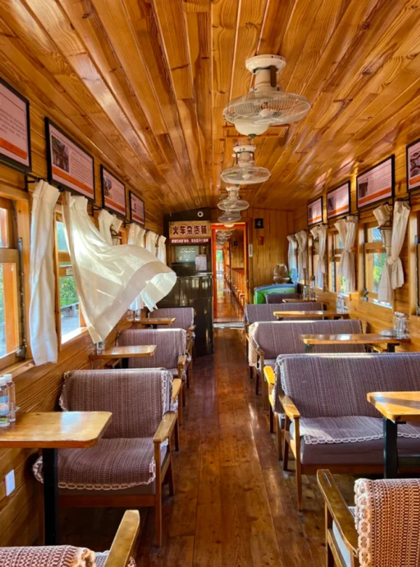 建水小火車復古車廂內部景觀，木質天花板與牆面營造溫暖氛圍，兩側窗戶配有白色窗簾與木桌座椅，車廂內整齊排列的座位與吊扇呈現懷舊鐵道風格，展現建水小火車獨特的歷史與觀光魅力