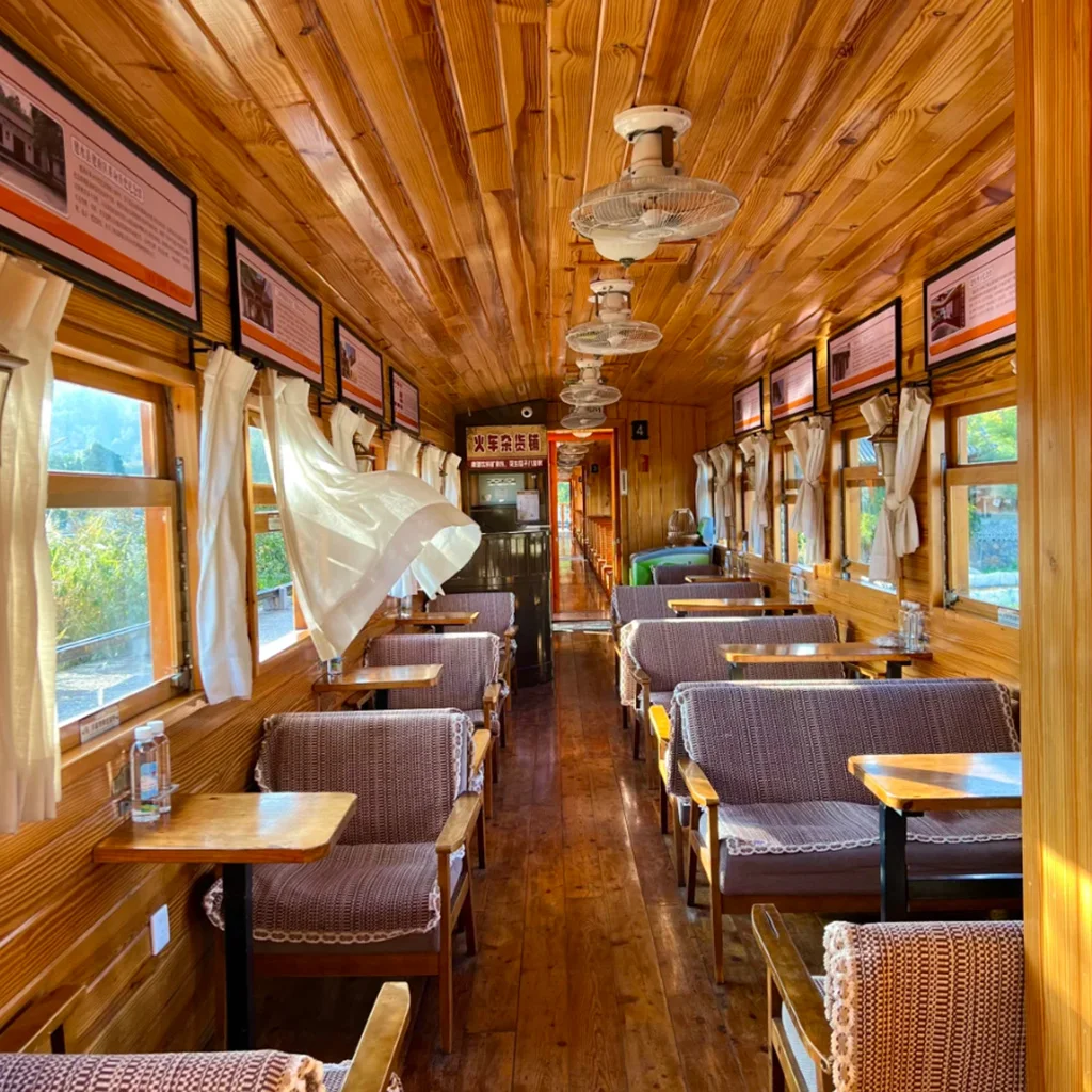 建水小火車復古車廂內部景觀，木質天花板與牆面營造溫暖氛圍，兩側窗戶配有白色窗簾與木桌座椅，車廂內整齊排列的座位與吊扇呈現懷舊鐵道風格，展現建水小火車獨特的歷史與觀光魅力