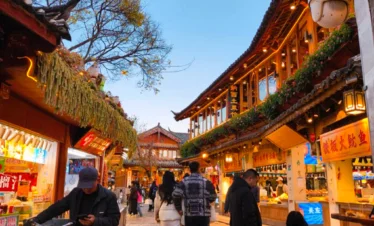 雲南著名歷史景區 麗江古城 的傍晚街景。石板路兩側是傳統木質建築與商店，屋簷掛著燈飾與招牌。遊客與當地居民在古街上漫步購物，街道燈光溫暖，營造出熱鬧又富有文化氣息的古鎮氛圍，展現麗江古城獨特的旅遊魅力