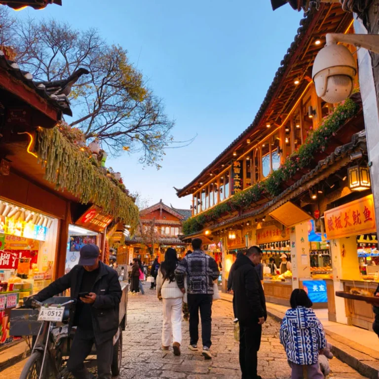 雲南著名歷史景區 麗江古城 的傍晚街景。石板路兩側是傳統木質建築與商店，屋簷掛著燈飾與招牌。遊客與當地居民在古街上漫步購物，街道燈光溫暖，營造出熱鬧又富有文化氣息的古鎮氛圍，展現麗江古城獨特的旅遊魅力
